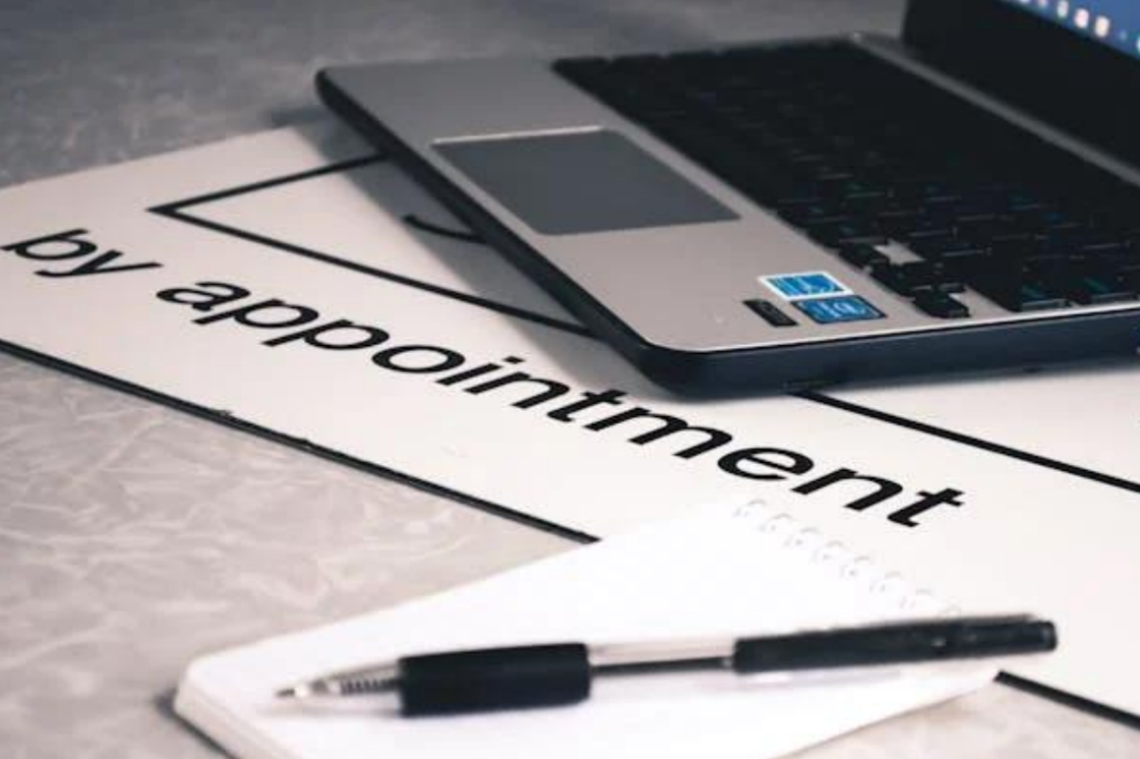 A laptop, notepad, and pen on a desk mat that reads ‘by appointment’.